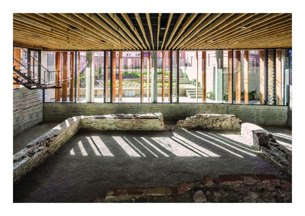 Innenansicht eines Raumes im Erdgeschoß mit Holzdecke; Ausgrabungsstätte mit Mauerresten; durchgehende Fensterfront mit Holzlamellen