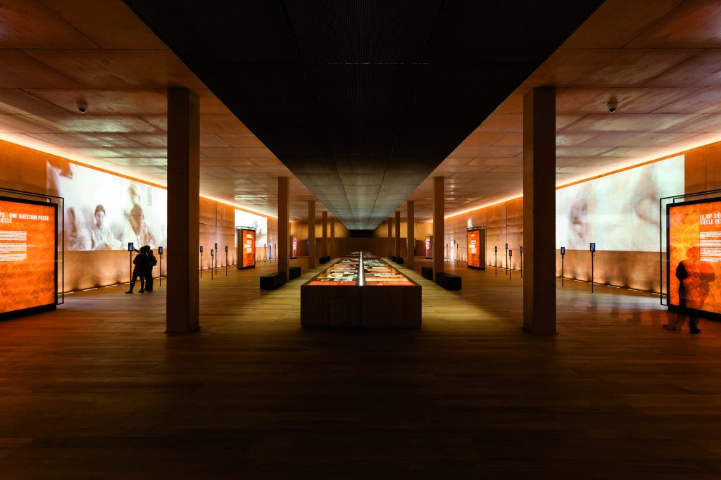 Innesansicht eines länglichen Ausstellungsraumes mit Säulen und einer langen Vitrine in der Mitte und Projektionen auf den Seitenwände