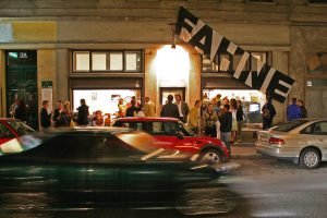 several people in front of a pub, some cars and a flag with the inscription ‘Fahne’ in front of it