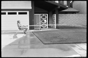 b/w photo of a man in an armchair spraying a meadow with a garden hose