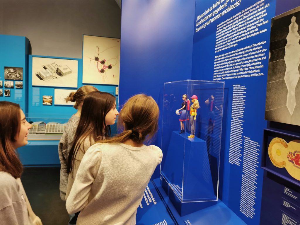 Four girls stand in front of a blue wall with texts and two Barbie dolls on display